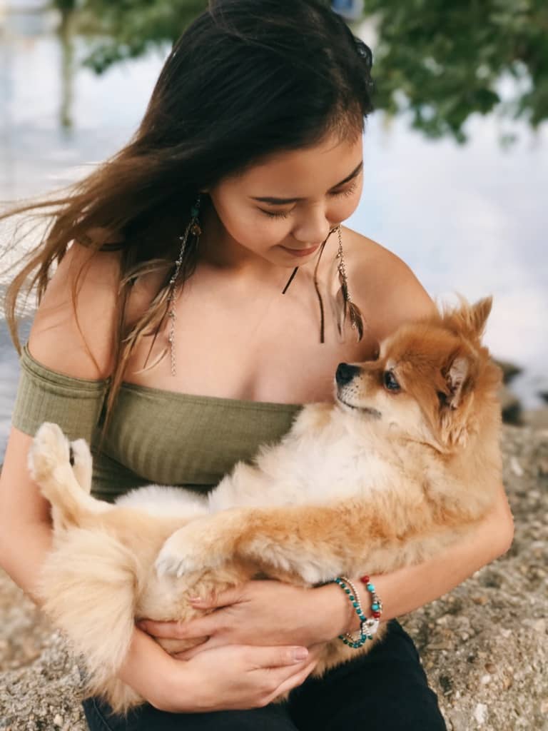 girl holding a golden pomeranian dog like a baby next to a lakefront during summer time