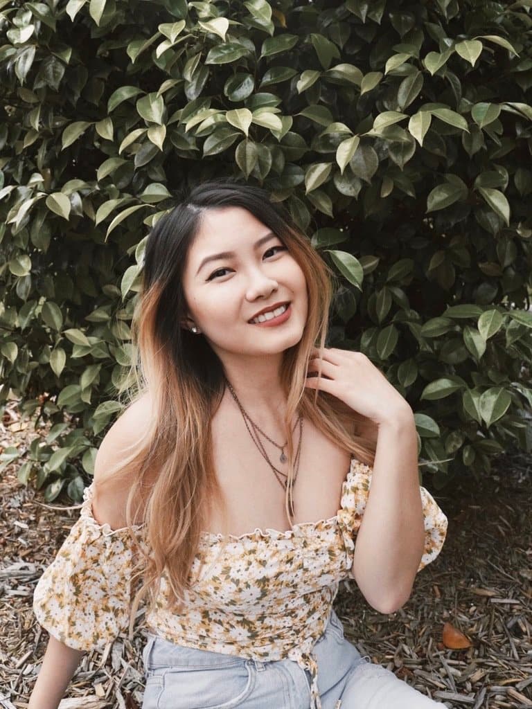 asian blonde girl sitting in front of green leaves bush in yellow off the shoulder top